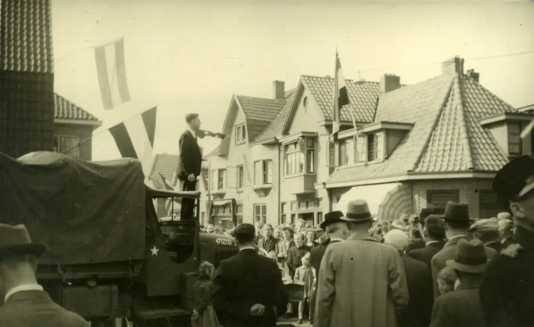 Oude foto van einde van Tweede Wereldoorlog in Borculo, een meneer spreekt in een drukke straat mensen toe vanaf het dak van een militaire auto